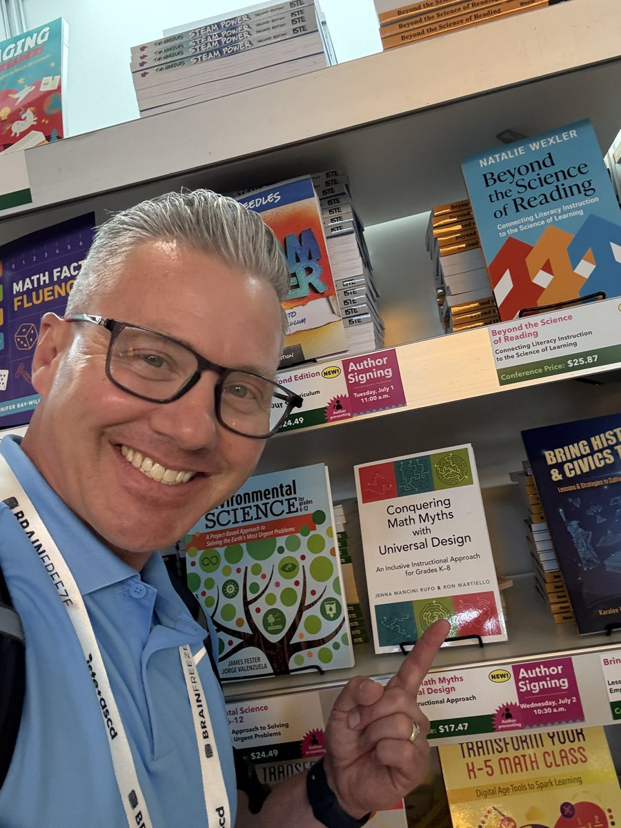 Ron Martiello, co-author of the book "Conquering Math Myths with Universal Design," smiling and pointing directly at his book displayed on a bookshelf at a conference. Other educational books are visible around him.